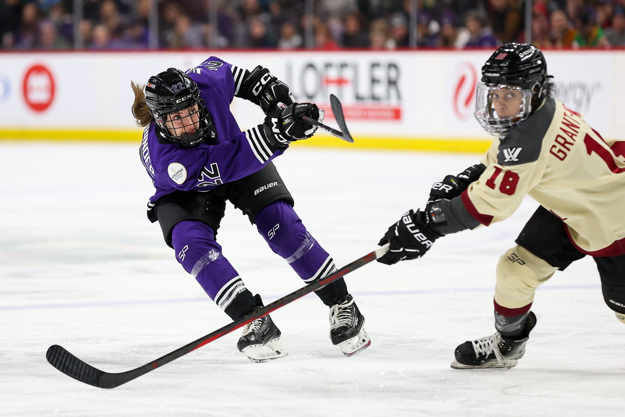 Natalie Buchbinder takes a shot against Montrรฉal as Mikyla Grant-Mentis defends. Buchbinder is hunched over with her stick raised in a follow-through. Grant-Mentis is hunched over with her stick on the ice outstretched to try to defend. Buchbinder is in purple, Grant-Mentis is cream.