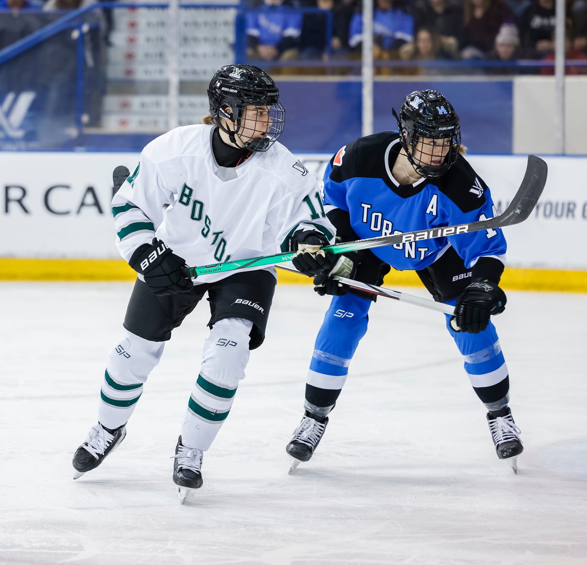 Alina Mรผller and Renata Fast hunt the puck. Mรผller is in white and is skating with her stick at waist-height. Fast is in blue and skating a bit hunched over with her stick also at wasit-heigh.