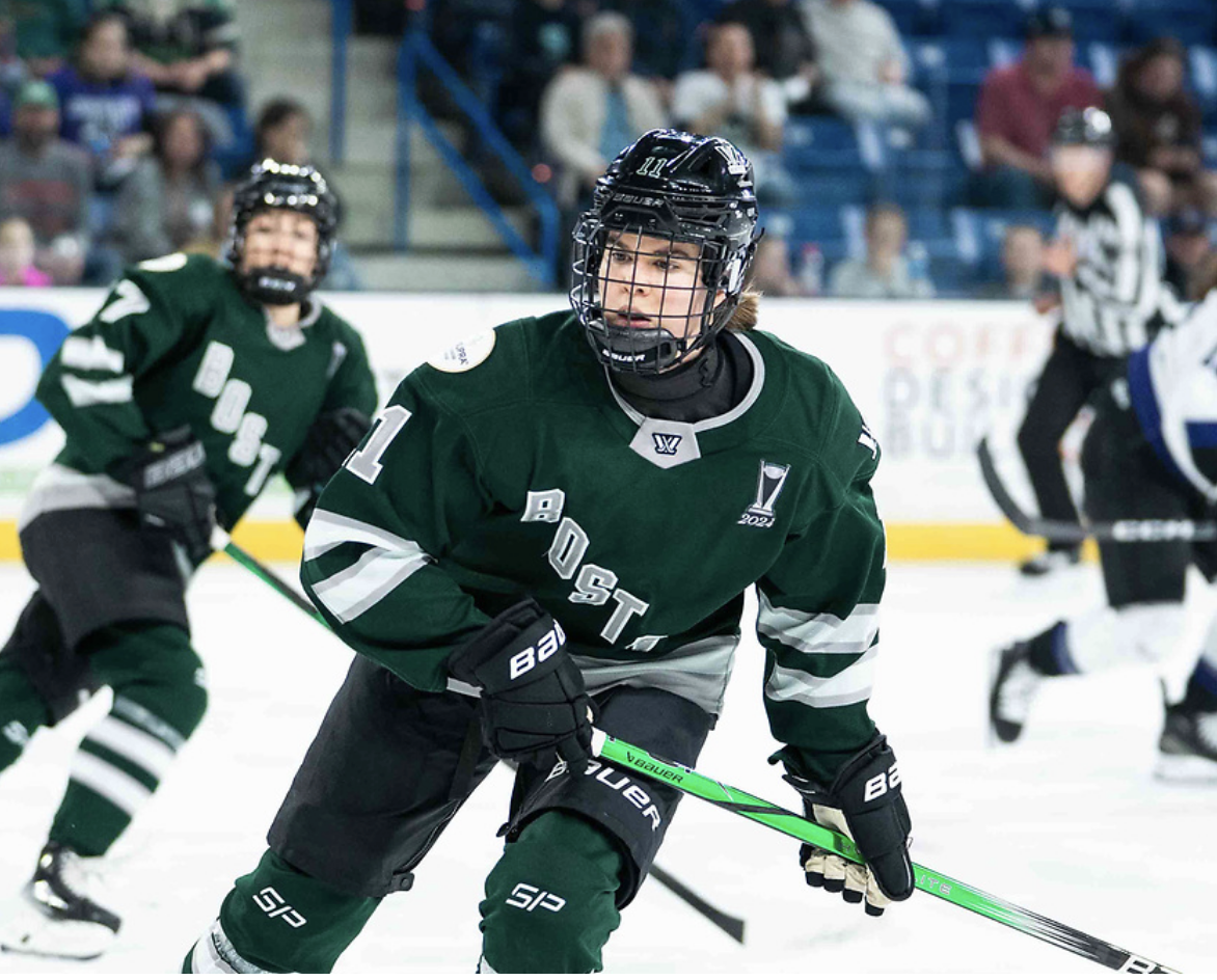 Müller watches a play develop while in pursuit of the puck. She is slightly crouched while skating, and wearing a green home uniform.