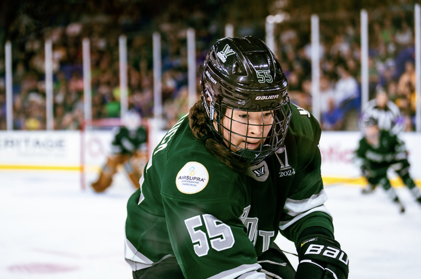 Babstock looks down at the out-of-frame puck, trying to scoop it out from along the boards. She is skating up ice wearing a green home jersey.