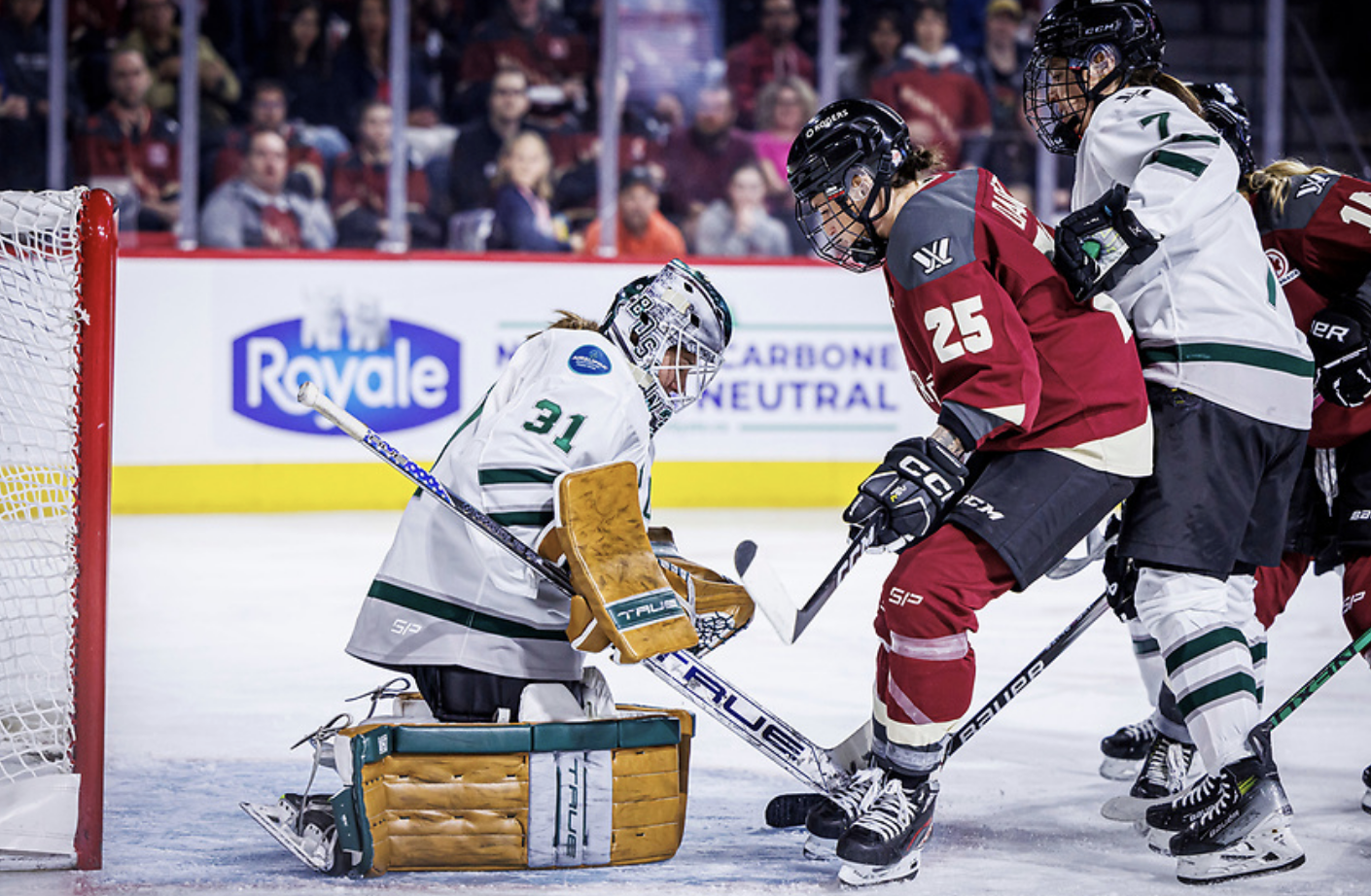 Frankel is on her knees, leaning slightly forward to make a save. Montrรฉal's Mรฉlodie Daoust is standing right in front of her, while Sidney Morin pushes her from behind. The Boston players are wearing white, while Daoust is wearing maroon.