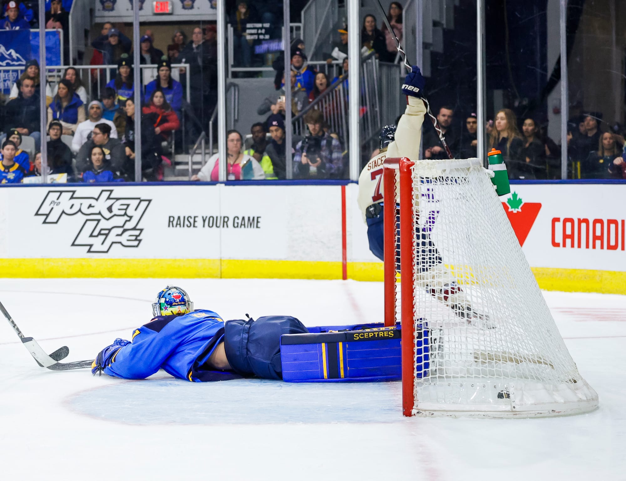 Kirk (wearing blue) lays flat on her stomach in the crease. In the background, Stacey (wearing cream) raises her stick in celebrating after scoring.