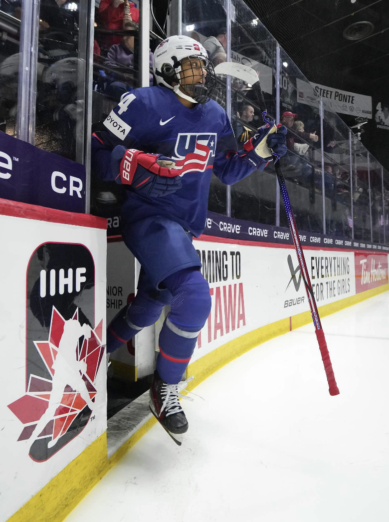 Edwards emerges onto the ice from an open door. She is looking straight ahead with her stick in her left hand, and wearing a blue USA uniform.