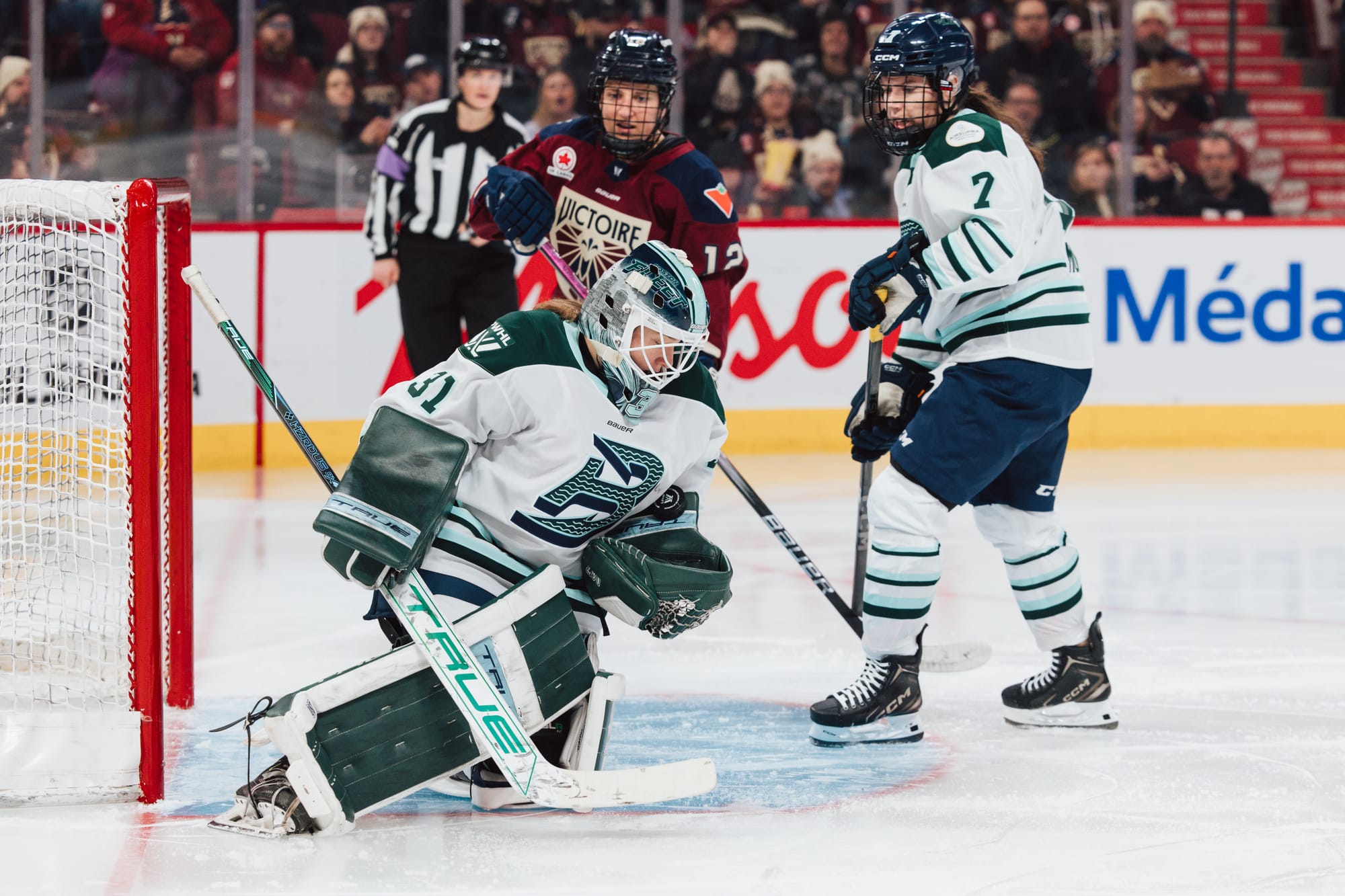 Frankel is kneeling on the ground, looking down at her glove as she cradles the puck. She is wearing a white away uniform.