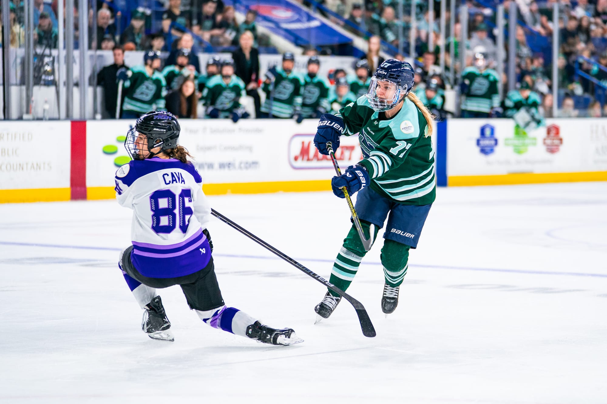 Tapani follows through with her stick after playing the puck, which is now out of frame. Minnesota's Michela Cava is on one knee after trying to block the play. Tapani is wearing a green home uniform, while Cava is in white.
