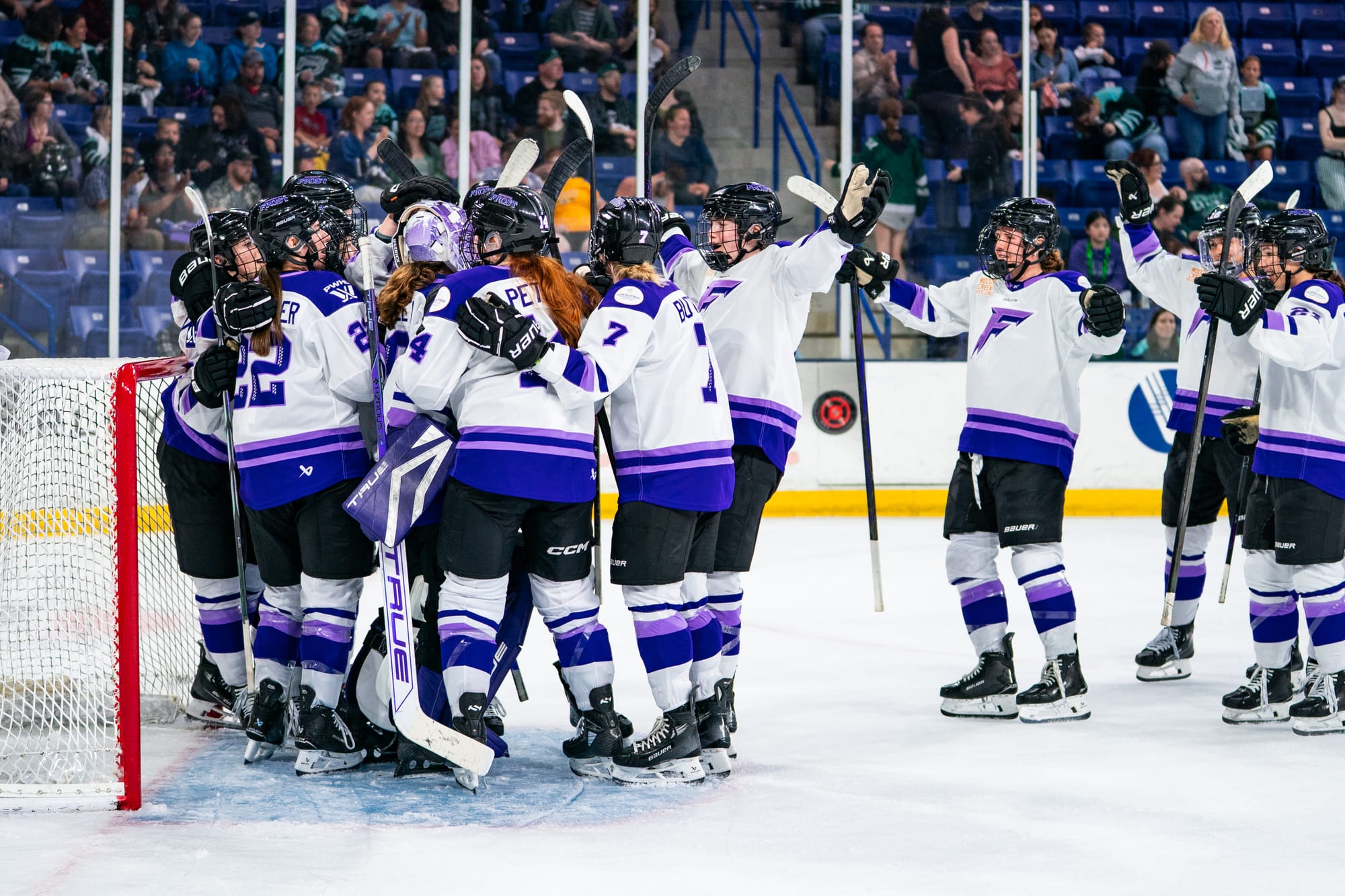 Minnesota players celebrate their win with a big group hug in Hensley's crease. They are all wearing white uniforms.