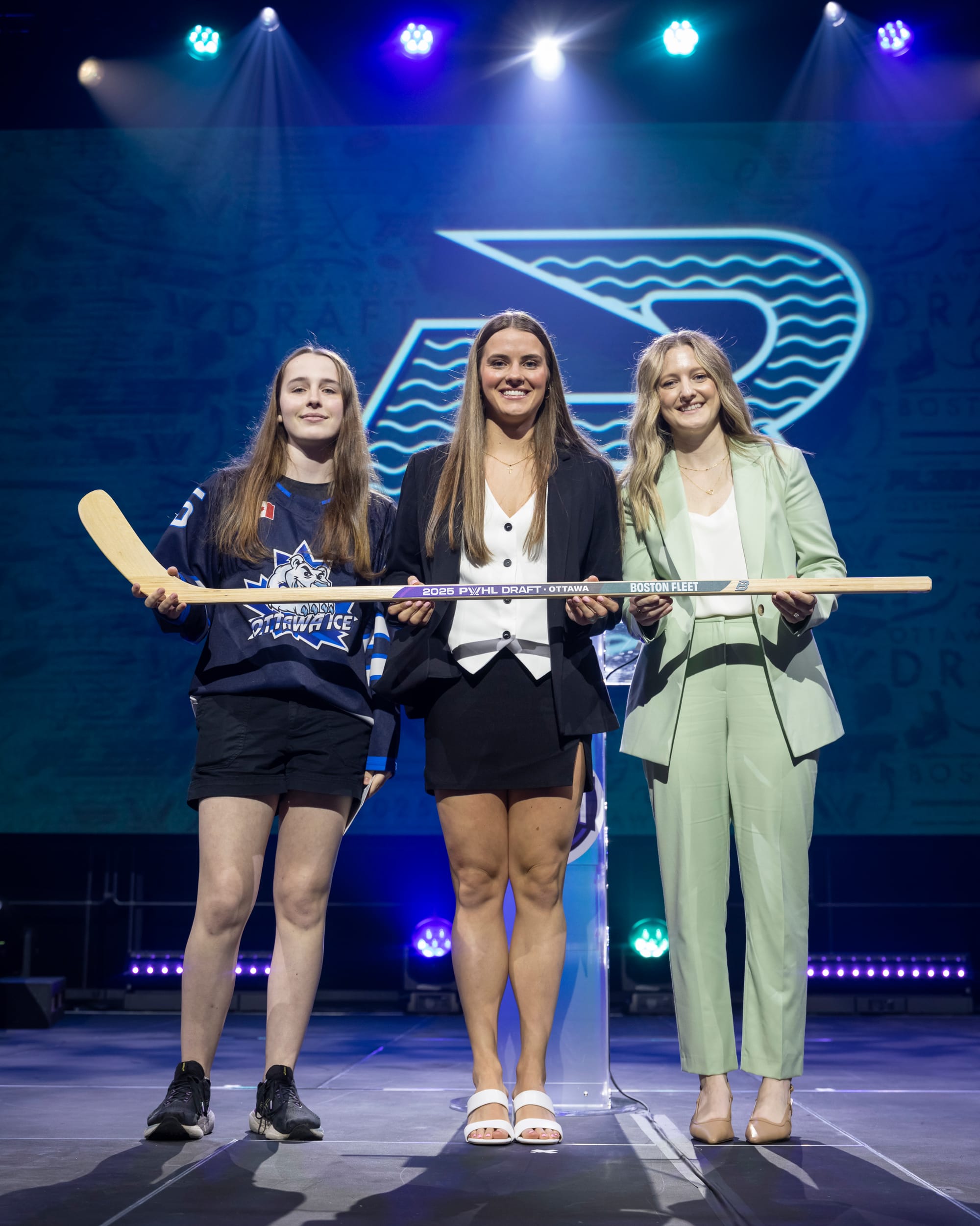 From left to right: a youth hockey player, Brengman, and Marmer smile on stage while holding a wooden stick that says "2025 PWHL Draft Ottawa- Boston Fleet."