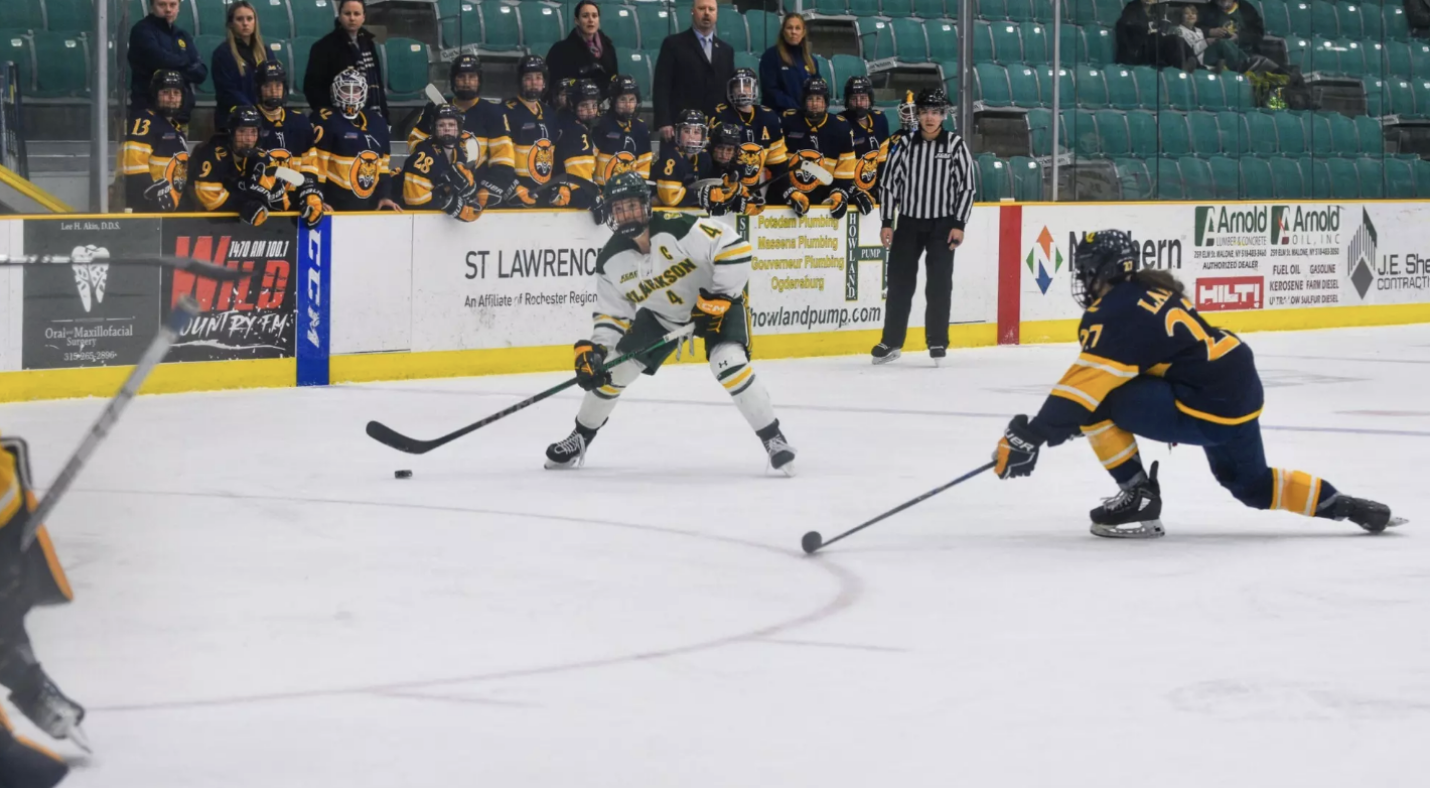 Winn skates with the puck in her team's offensive zone. She is wearing a white home uniform.