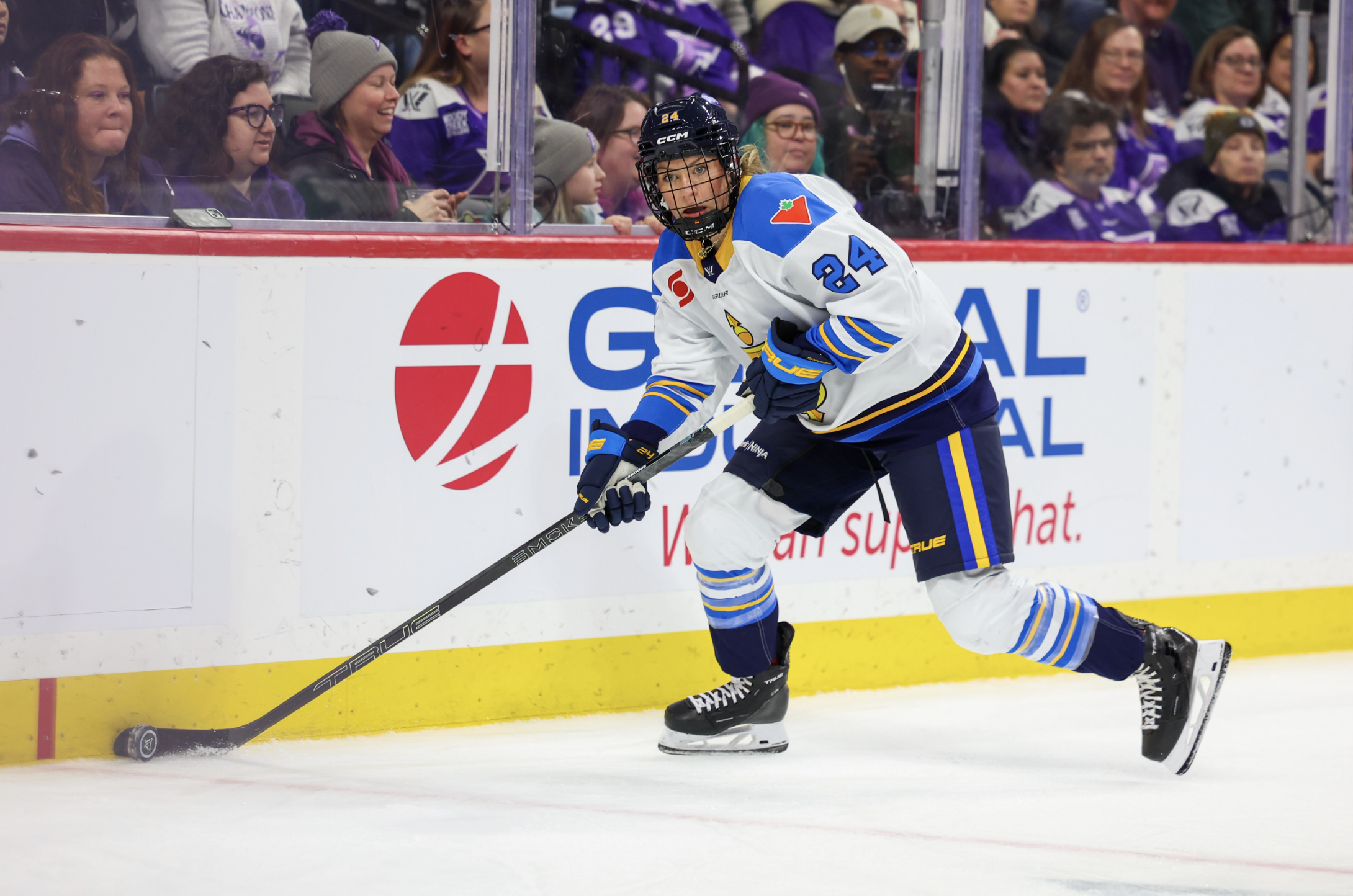 Spooners skates with the puck during their season opener. She is along the boards, wearing a white away uniform. 
