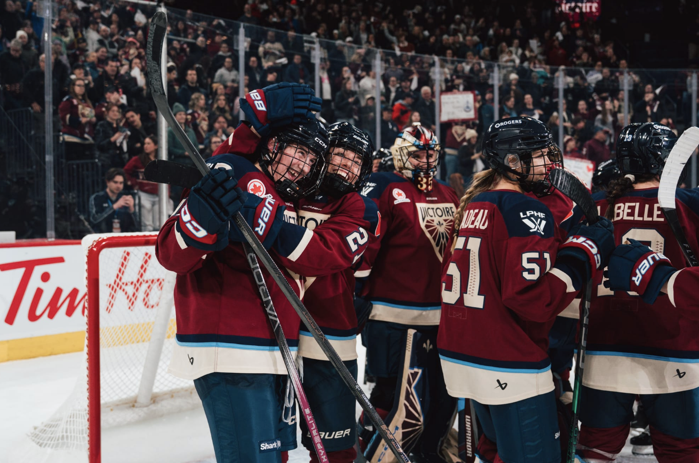 The Victoire celebrate with hugs around Ann-Renée Desbiens. They are all wearing maroon home uniforms.