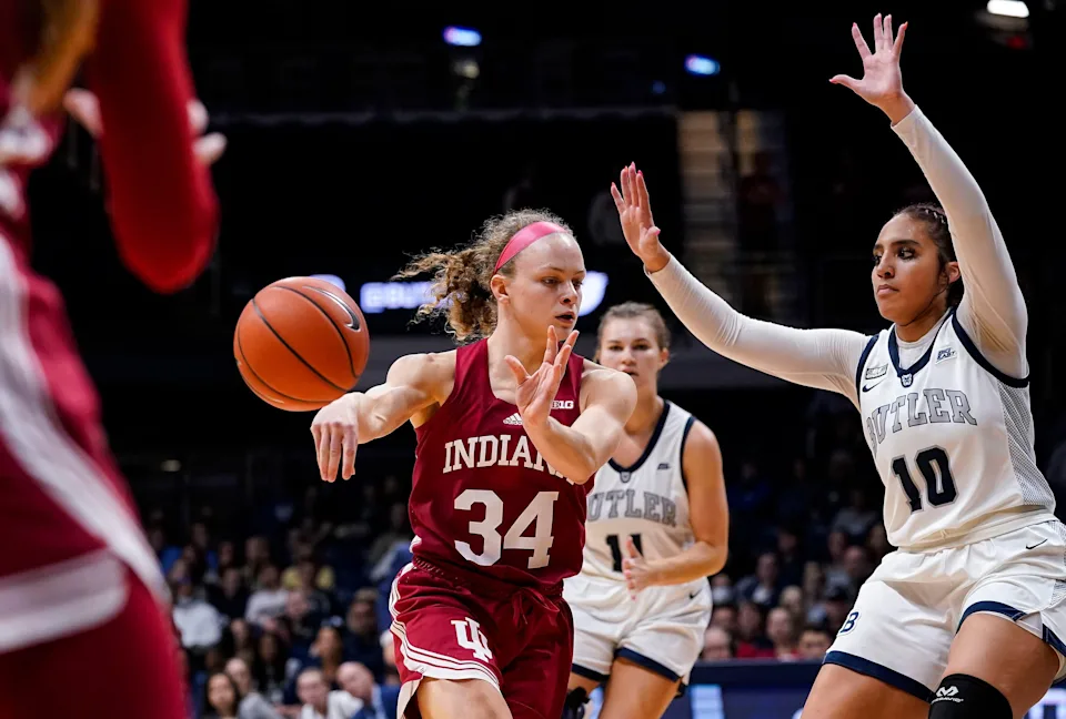 Grace Berger holds her follow-through on a no-look kick-out pass to a spot-up shooter whose hands are out to receive the ball in the foreground, while a defender has her hands up, trying to stay in front of Berger.