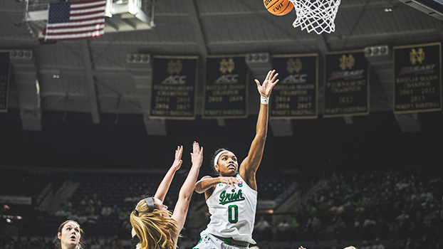 Maya Dodson, mid-air, watches with an outstretched arm as the ball she's laid-up goes up towards the hoop. A defender is grounded beside her, arms up to try to defend in vain, while another defender watches on.