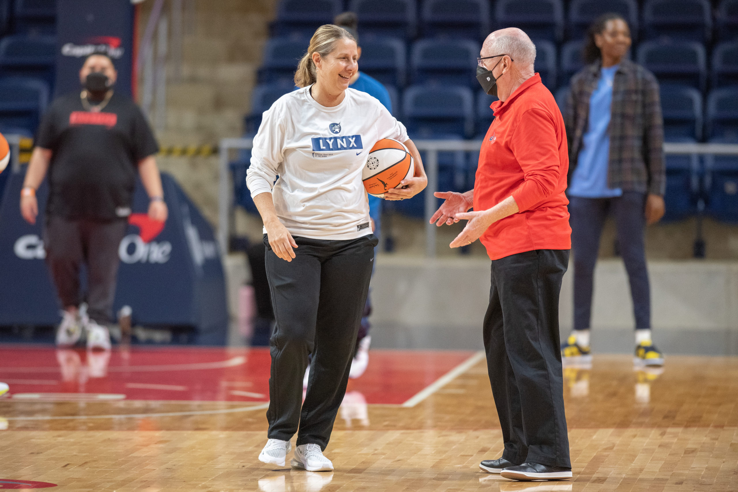 Minnesota Lynx head coach/general manager Cheryl Reeve stands near midcourt holding a ball under her arm and laughing to Washington Mystics head coach/general manager Mike Thibault, who stands beside her.