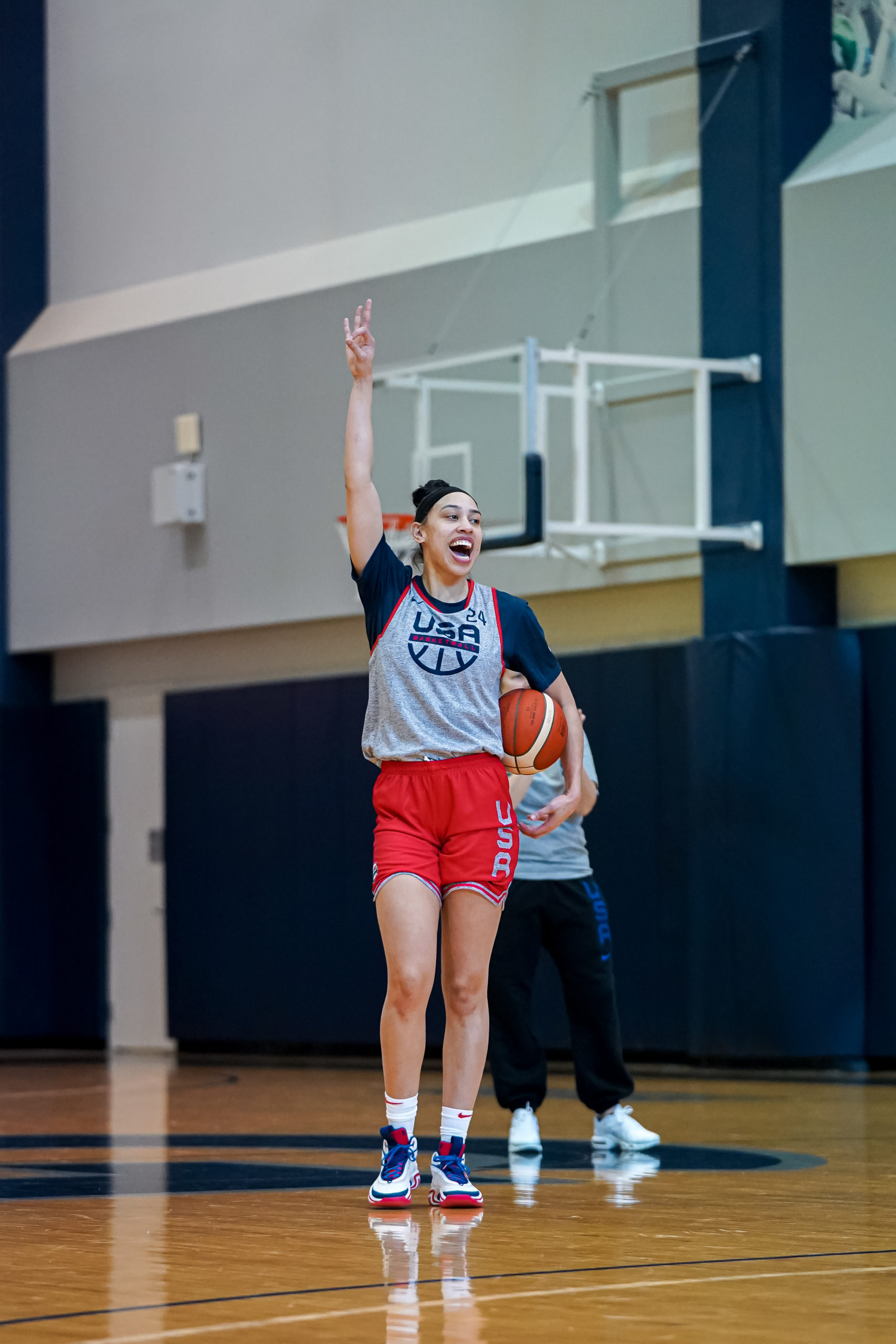 Dearica Hamby, wearing a USA practice pinnie, raises her right arm in the air while holding a basketball in her left.