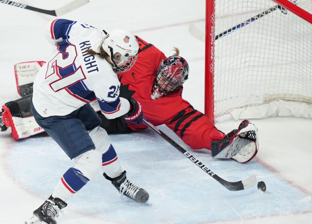Team USA player Hilary Knight, wearing a red, white and blue uniform attempts a shot close to the net on Canada's goaltender, who is wearing a red and black uniform and diving to attempt to stop the goal.