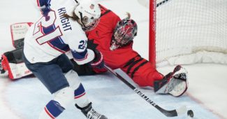 Team USA player Hilary Knight, wearing a red, white and blue uniform attempts a shot close to the net on Canada's goaltender, who is wearing a red and black uniform and diving to attempt to stop the goal.