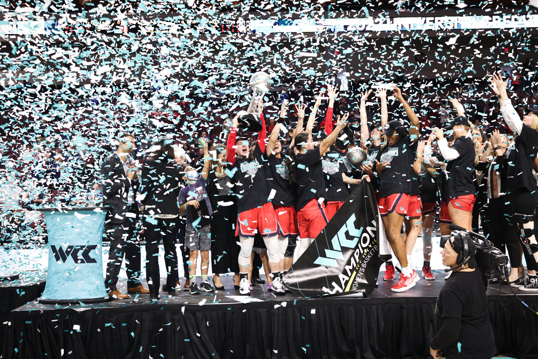 The Gonzaga Bulldogs women's basketball team celebrate after winning the WCC tournament, holding the trophy high as blue and white confetti falls around them
