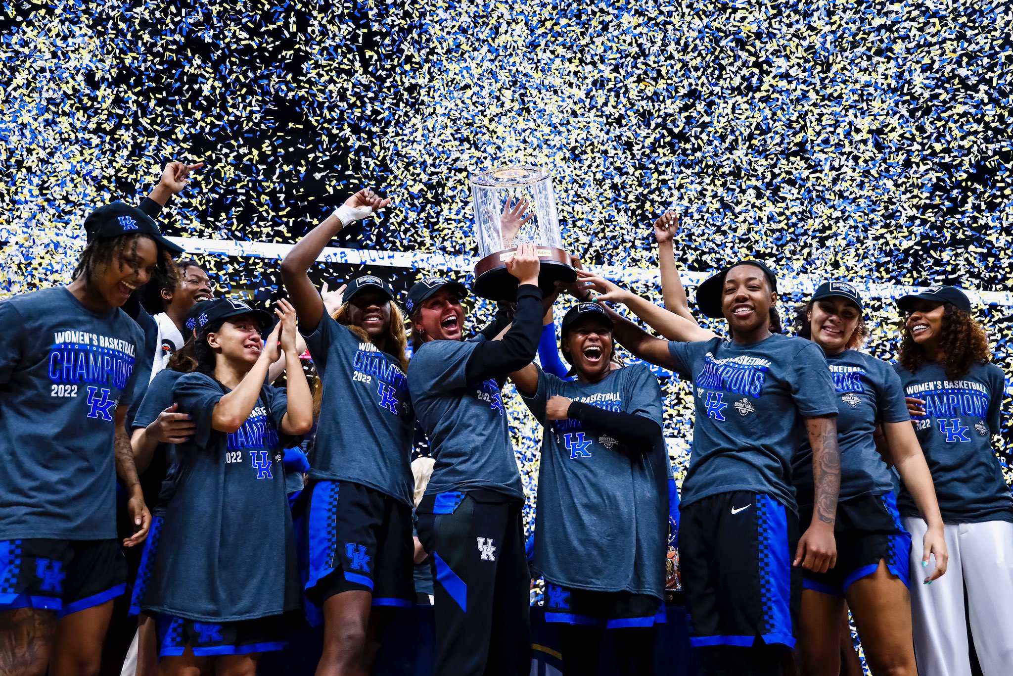 Kentucky Wildcats smile and cheer in championship shirts and hats while holding up the SEC trophy as blue and white confetti showers them