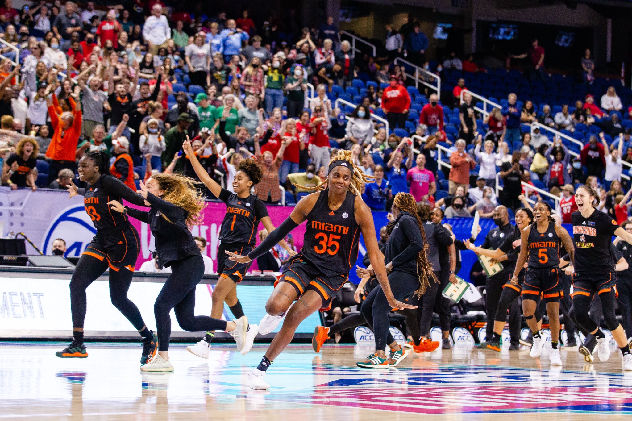 Miami players rush to halfcourt to celebrate while Naomi Mbandu does an airplane celebration and fans in the stands in the background celebrate wildly