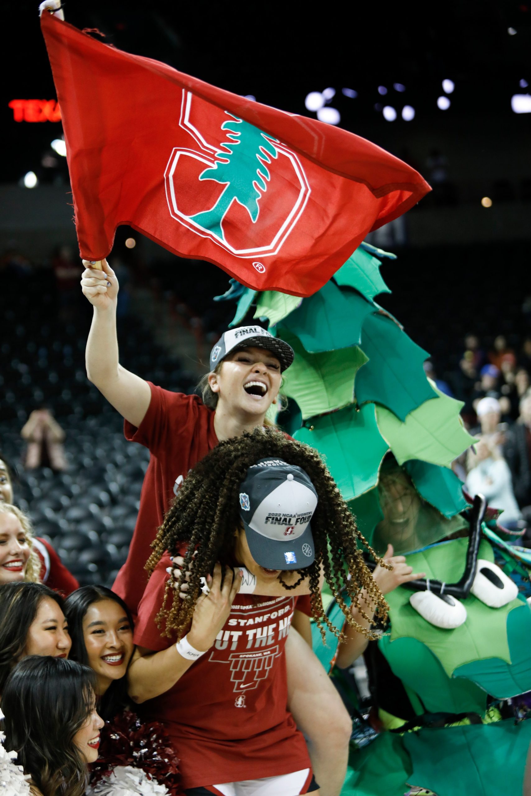 A Stanford player smiles widely while riding on Haley Jones' back and waving a Stanford flag with one hand. Some cheerleaders pose next to them for a photo, while on their other side, the Cardinal mascot smiles and gives a thumbs-up.