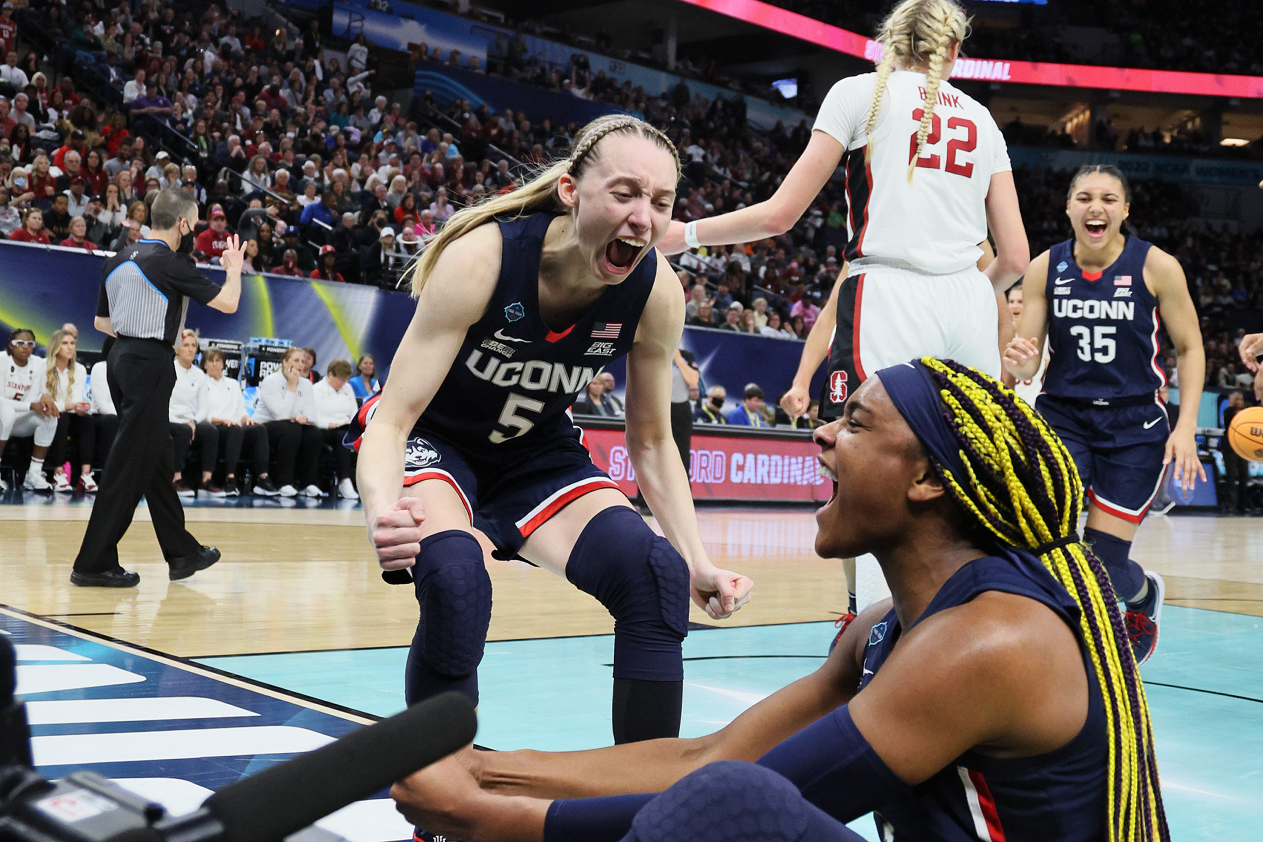 Paige Bueckers, crouching, and Aaliyah Edwards, sitting on the ground, yell in celebration, while Azzi Fudd runs in, smiling, from the background to celebrate. Stanford Cardinal Cameron Brink walks away from Bueckers and Edwards.