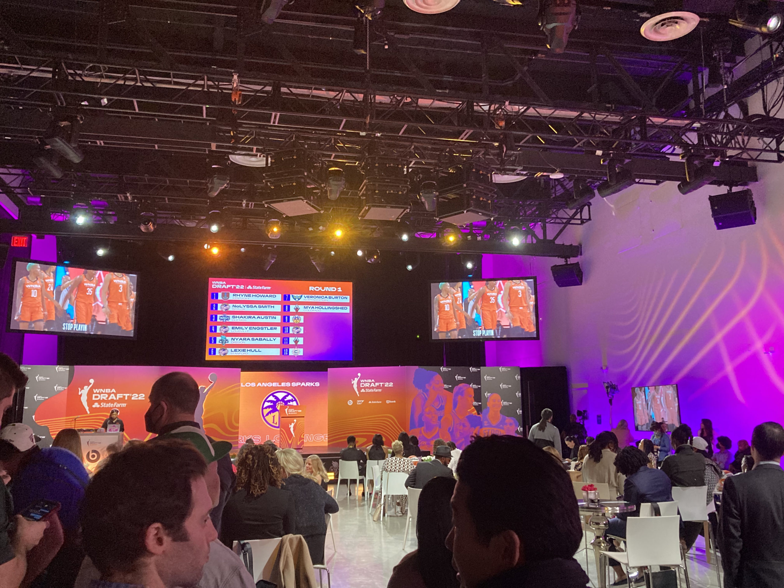 A picture taken from the 2022 WNBA Draft, looking out across tables with potential draftees and their friends and families, and a stage with an orange WNBA branded podium and screens showing the night's draft picks so far.