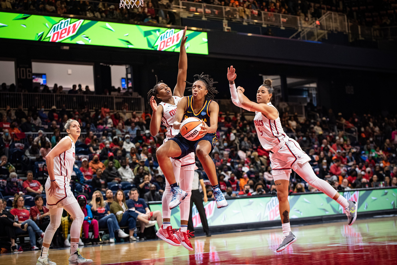 Indiana Fever combo guard Kelsey Mitchell holds the ball with both hands while her legs are pulled up in midair, as two Washington Mystics defenders stand watching behind her