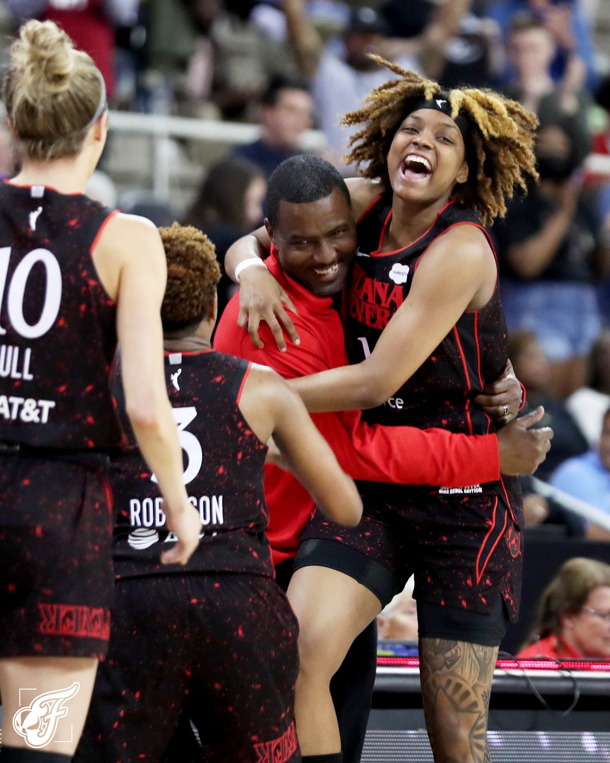 Fever head coach Carlos Knox embraces Fever forward NaLyssa Smith, both are smiling. Fever guards Lexie Hull and Danielle Robinson walk towards Knox and Smith.