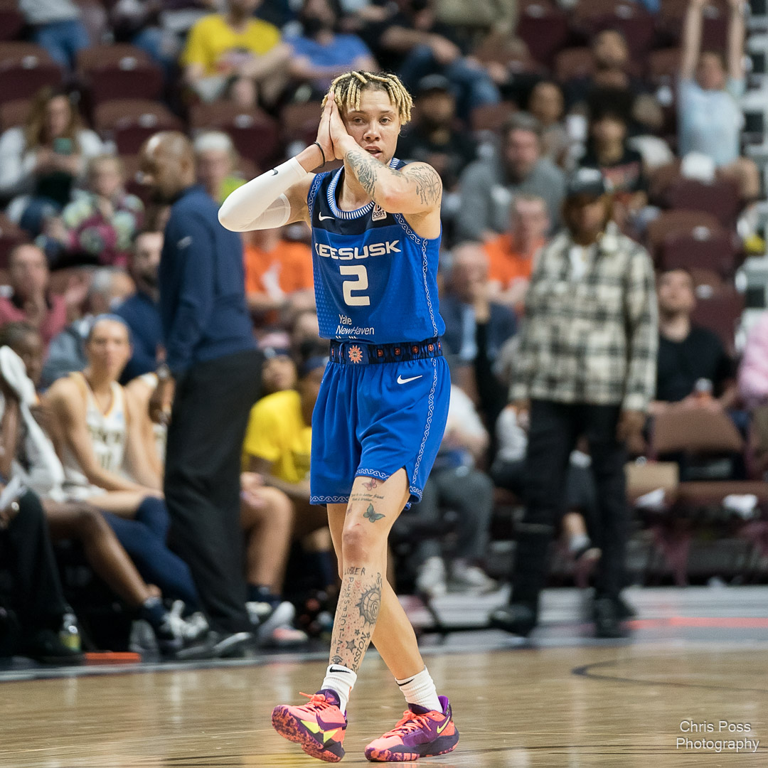 Connecticut Sun point guard Natisha Hiedeman walks down the court with her palms together against her cheek, making the "sleep" motion. The background shows some of the crowd and the Indiana Fever bench, but it's blurred out.
