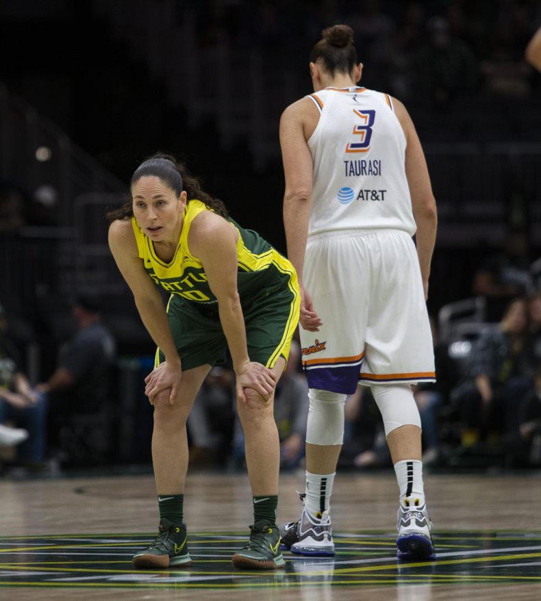 Seattle Storm point guard Sue Bird hunches over with her hands on her knees, facing slightly towards the camera, while Phoenix Mercury wing <a rel=