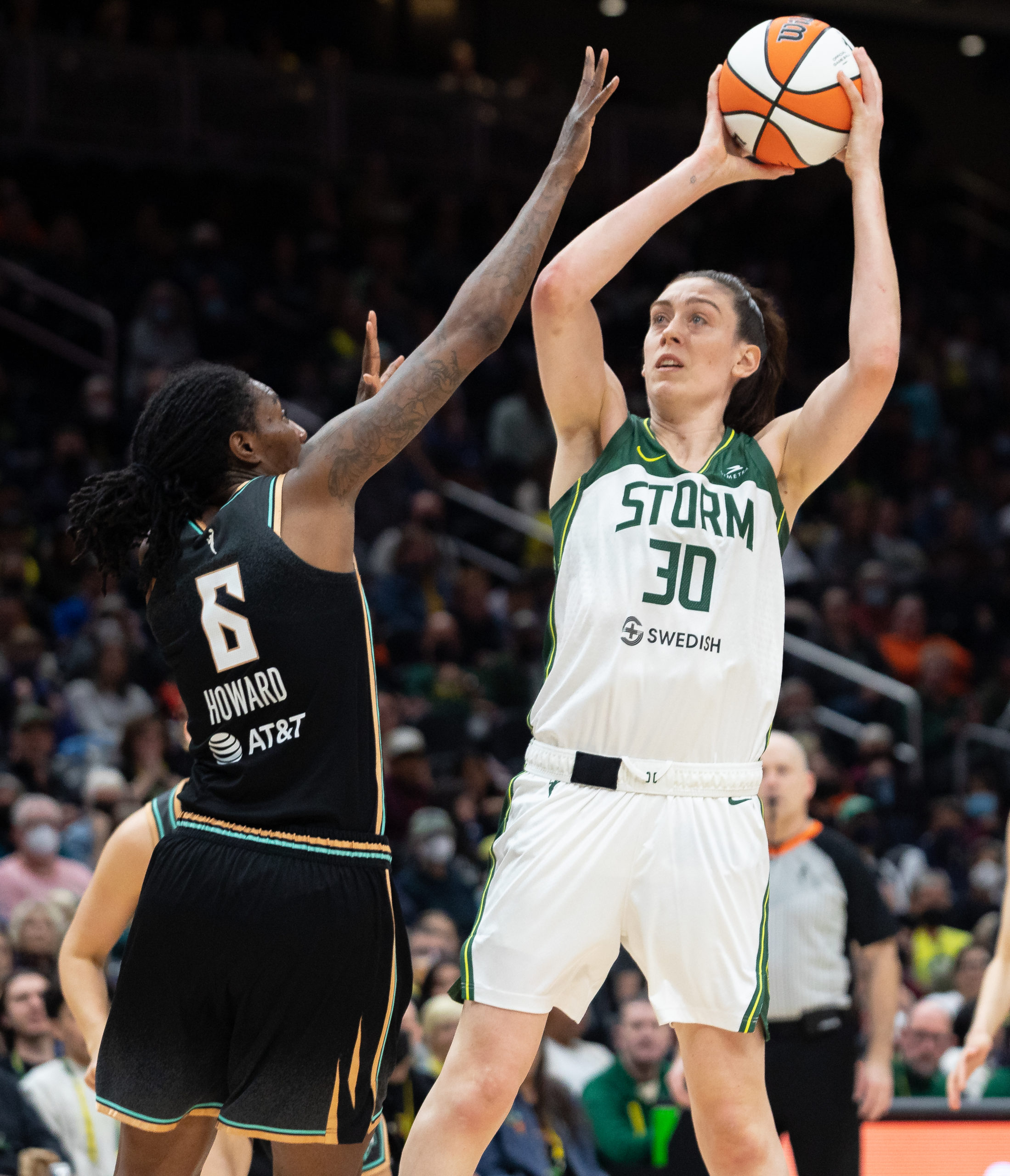 Seattle Storm big wing Breanna Stewart leans back with the ball high above her head as she begins to shoot a fadeaway jumper, while New York Liberty big Natasha Howard leans toward her with an arm outstretched to try to block the shot.