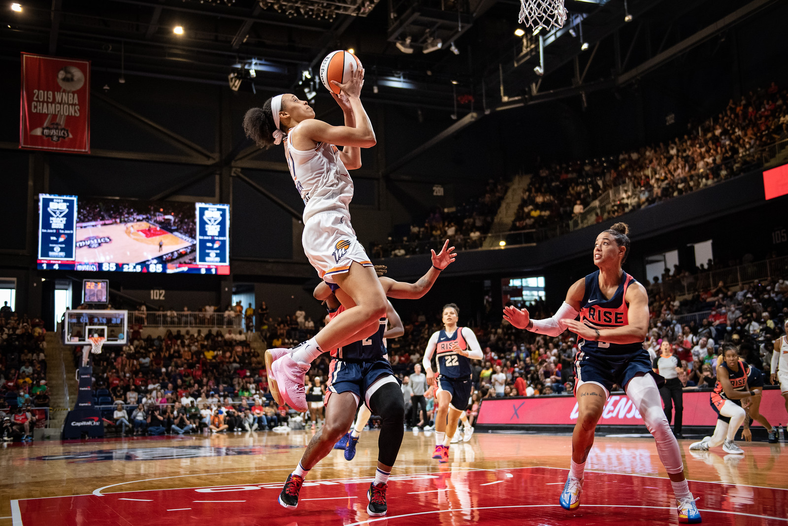 Phoenix Mercury big Brianna Turner, in mid-air above the restricted area, holds the ball in front of her face while looking at the hoop. Washington Mystics defenders stand behind her looking very concerned.