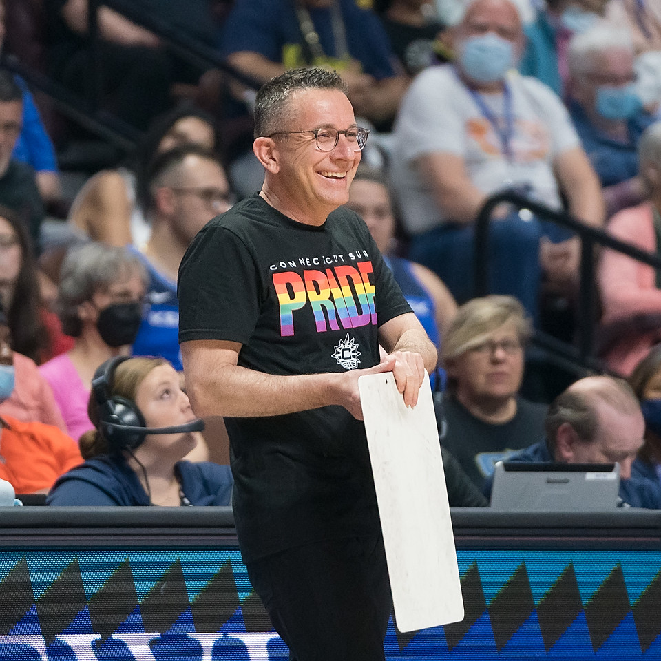 Connecticut Sun head coach Curt Miller stands in front of the scorer's table, holding a clipboard and smiling while looking out at the court. He is wearing the Sun's Pride shirt.