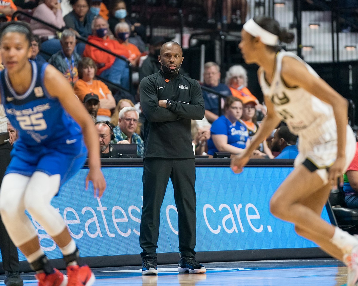 Chicago head coach James Wade stands in front of the scorer's table, looking towards the halfcourt, where an out-of-focus Sky combo forward Rebekah Gardner and Connecticut Sun big Alyssa Thomas stand
