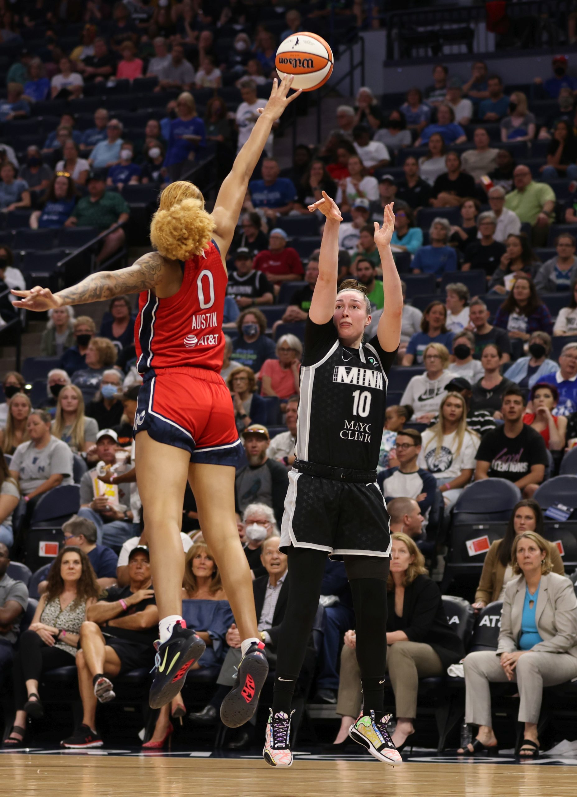 Minnesota Lynx combo forward Jessica Shepard, mid-air, holds her follow-through while watching the ball travel towards the hoop, as Washington Mystics center Shakira Austin jumps towards her with an outstretched arm in an attempt to contest the shot