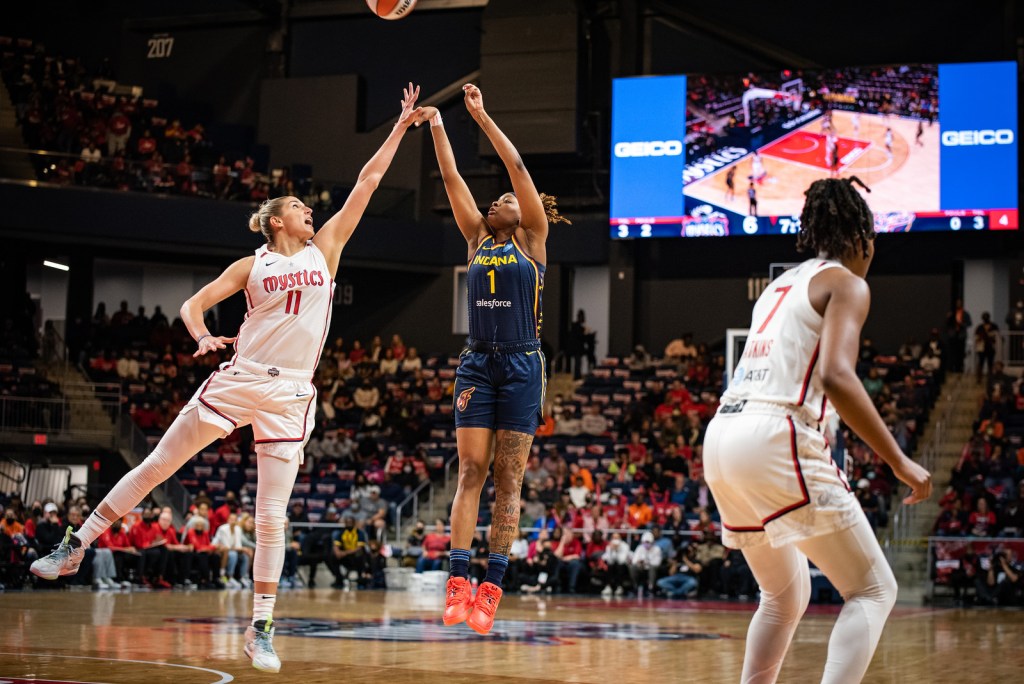 Indiana Fever big NaLyssa Smith descends from mid-air while holding her jumper form as the ball travels towards the rim, while Washington Mystics big Elena Delle Donne leans towards Smith in midair with an outstretched arm in a failed attempt to block the shot