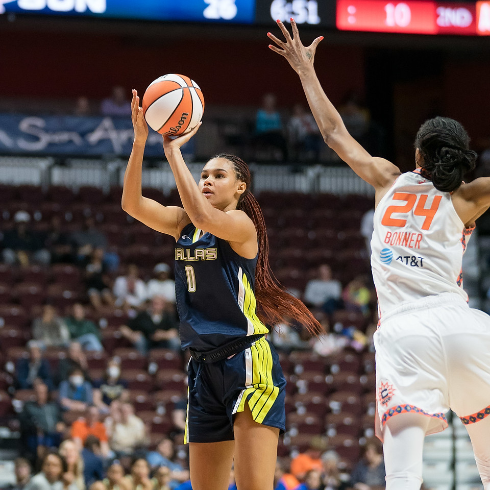 Dallas Wings big wing Satou Sabally has the ball out and above her head, as she releases a three-pointer, while Connecticut Sun big wing DeWanna Bonner lunges towards her with her arm outstretched in an attempt to contest the shot