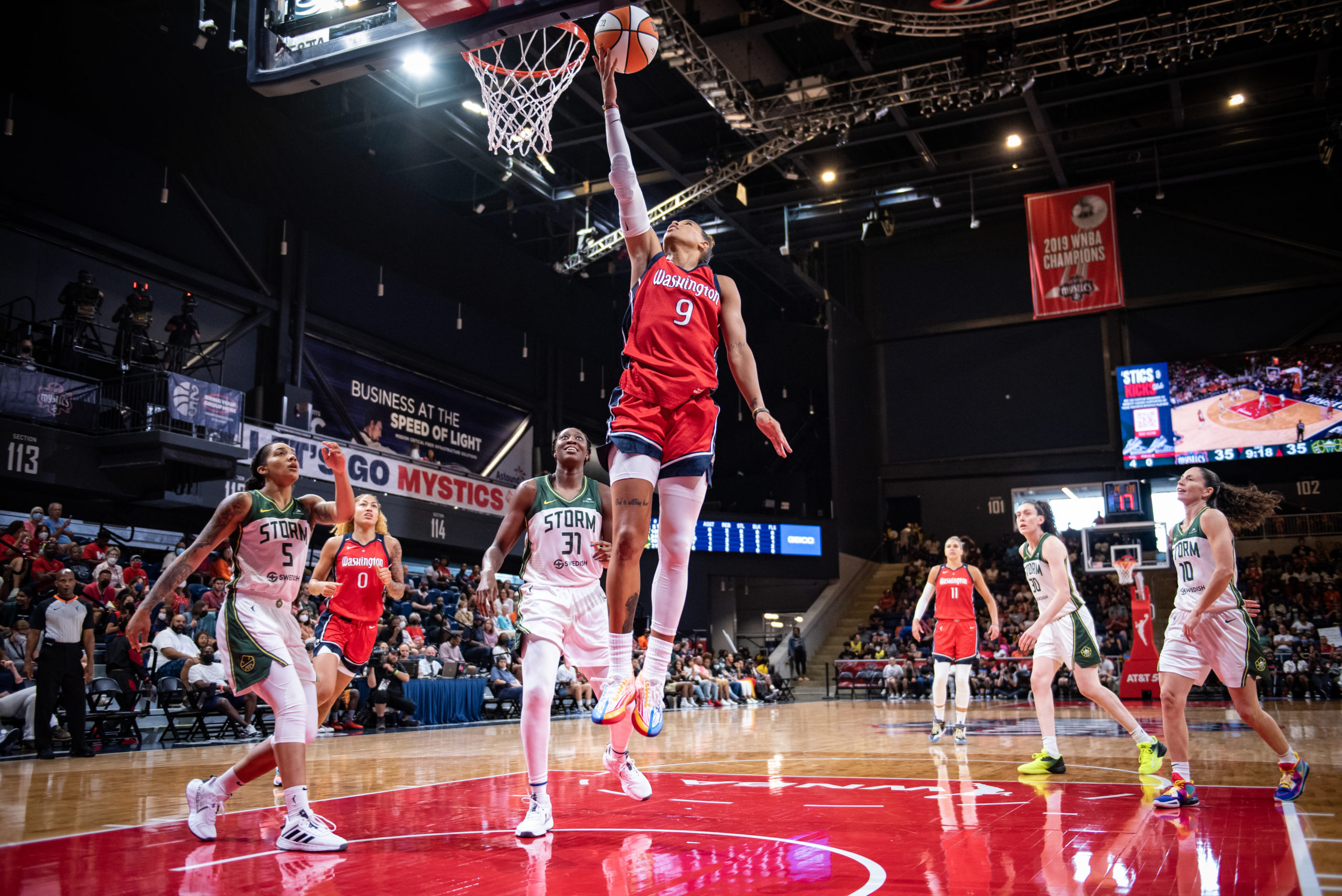 Mystics point guard Natasha Cloud (9) goes for a finger-roll layup. Storm center Tina Charles (31) and off-ball guard Gabby Williams (5) stand close behind, while point guard Sue Bird (10) and big win Breanna Stewart (30) stand at the top of the key. Mystics center Shakira Austin (0) stands behind Williams, while big Elena Delle Donne (11) stands behind Stewart.
