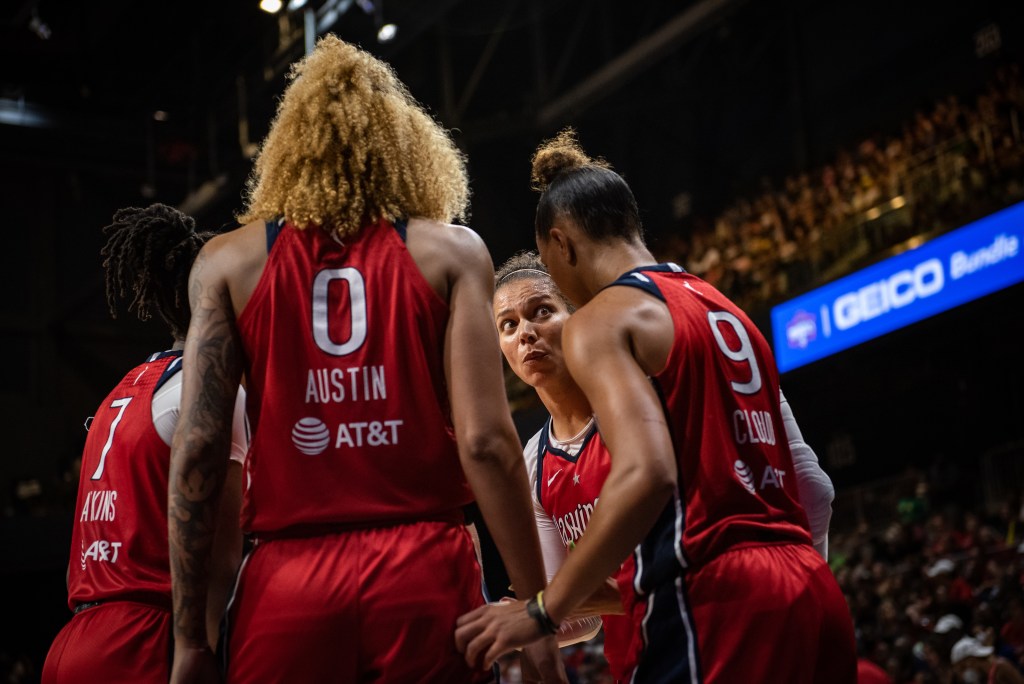 Mystics center Shakira Austin (0), point guard Natasha Cloud (9), off-ball guard Ariel Atkins (7), and off-ball guard Alysha Clark (22) huddle up. Clark makes a surprised face in Cloud's direction.