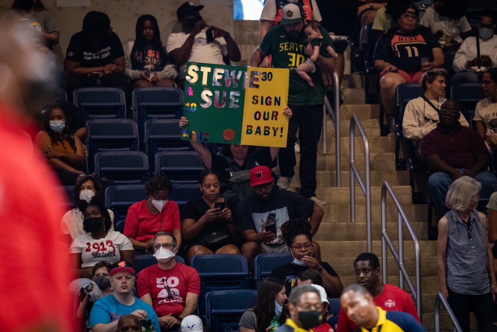 Fans hold a green and yellow sign asking Seattle Storm players Breanna Stewart and Sue Bird to "Sign our baby!" Lettering is in silver and black.