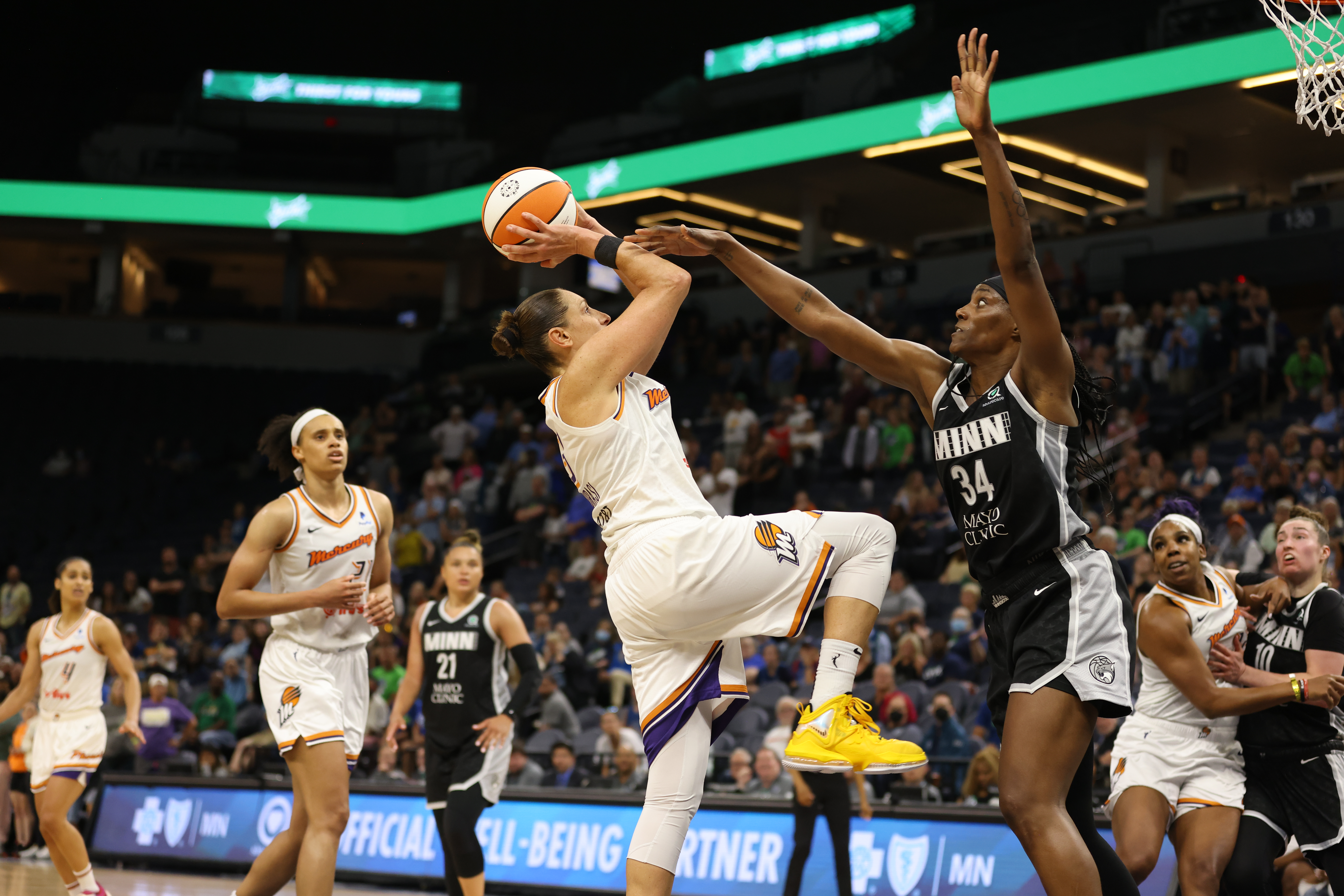 Phoenix Mercury combo guard Diana Taurasi holds the ball above her head as she attempts a short fadeaway jumper off of leg, while Minnesota Lynx center Sylvia Fowles stands in front of her with one arm straight up and one arm out towards the ball to contest the shot