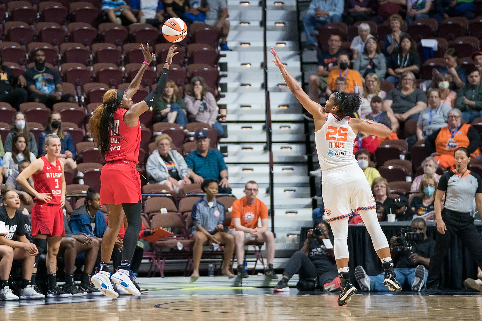 Atlanta Dream wing Rhyne Howard, slightly mid-air, releases a 3-pointer from the slot as Connecticut Sun big Alyssa Thomas lunges towards her with an arm outstretched to contest the shot.