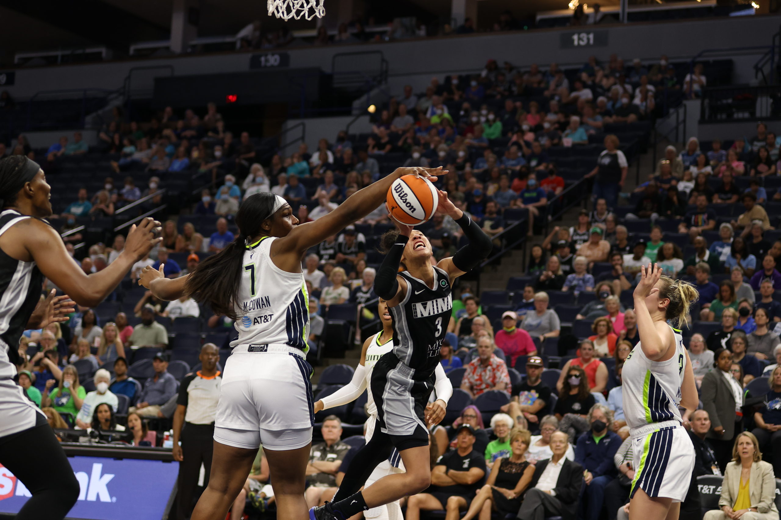 While standing firmly on the ground, Dallas Wings center Teaira McCowan holds her right arm out to her side and blocks a layup attempt from a mid-air, out-of-control Minnesota Lynx off-ball guard Aerial Powers