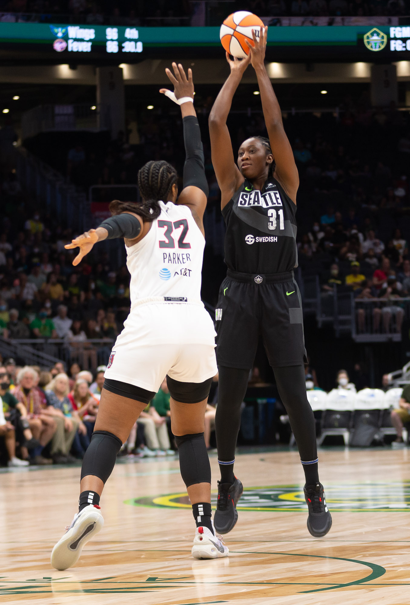 Seattle Storm center Tina Charles, mid-air, releases a jump shot off her fingertips from atop the key, as Atlanta Dream big Cheyenne Parker lunges toward her with an outstretched arm to contest the shot