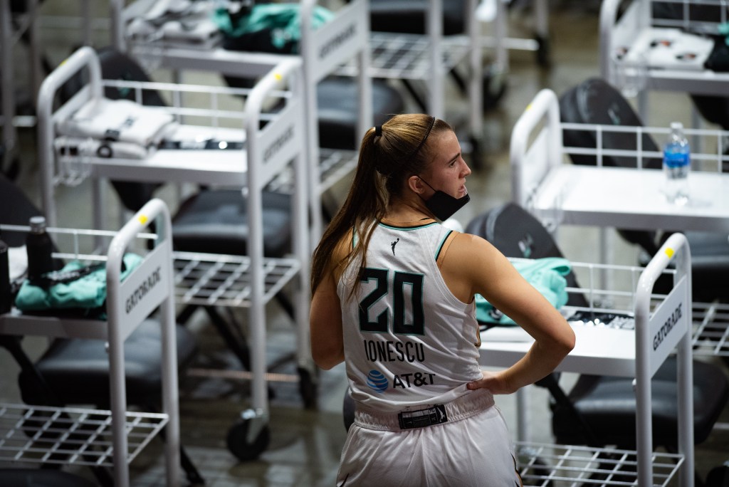 New York combo guard Sabrina Ionescu stands in front of a row of alternating chairs and carts that have towels and water bottles on them, looking to the side with a mask pulled down over her chin