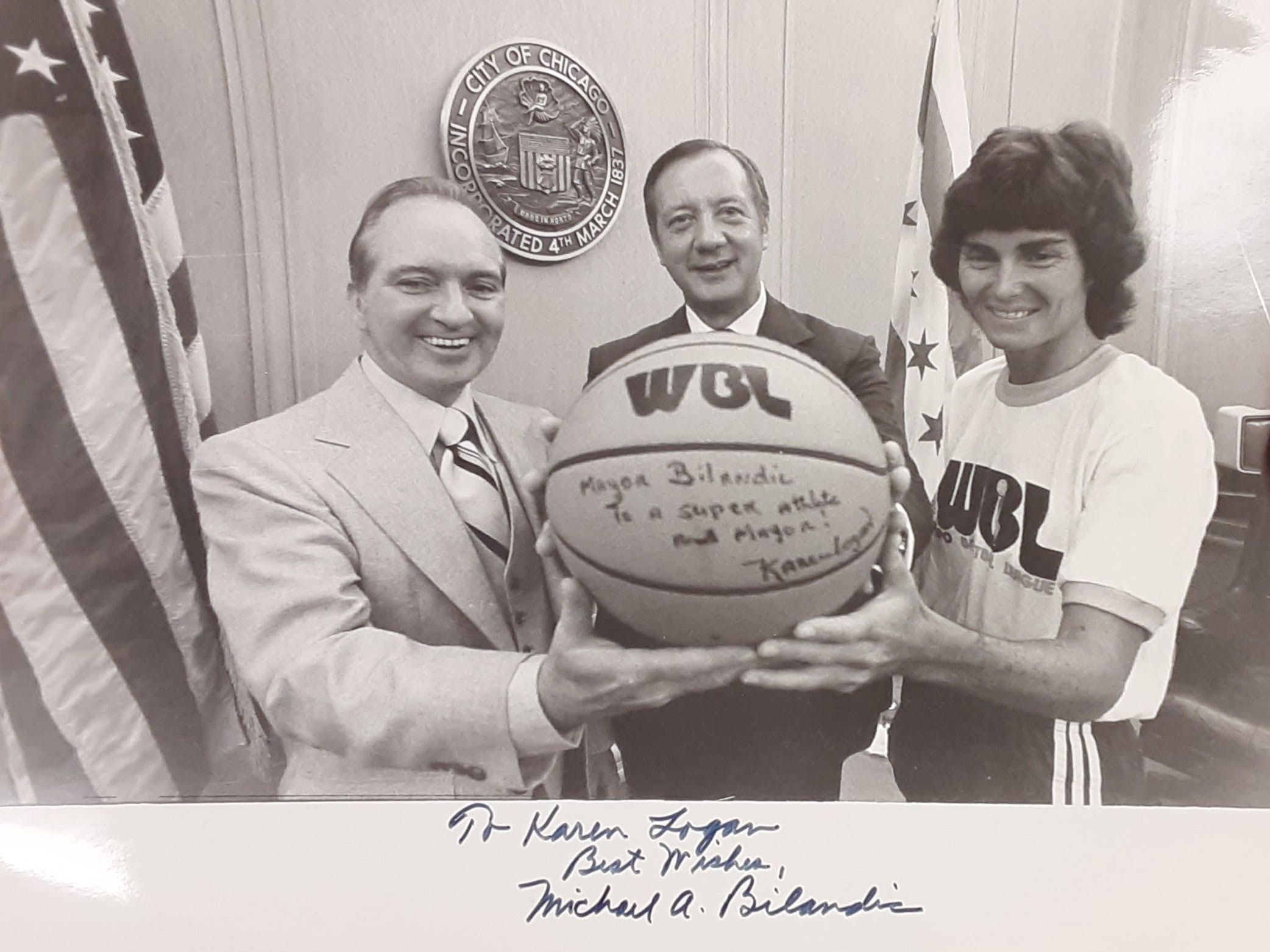 Karen Logan (right) and John Geraty (left), general manager of the Chicago Hustle, presents Chicago Mayor Michael A. Bilandic (center) with a signed WBL ball that reads "Mayor Bilandic, To a super athlete and Mayor -Karen Logan". The photo is signed by Bilandic with the message "To Karen Logan