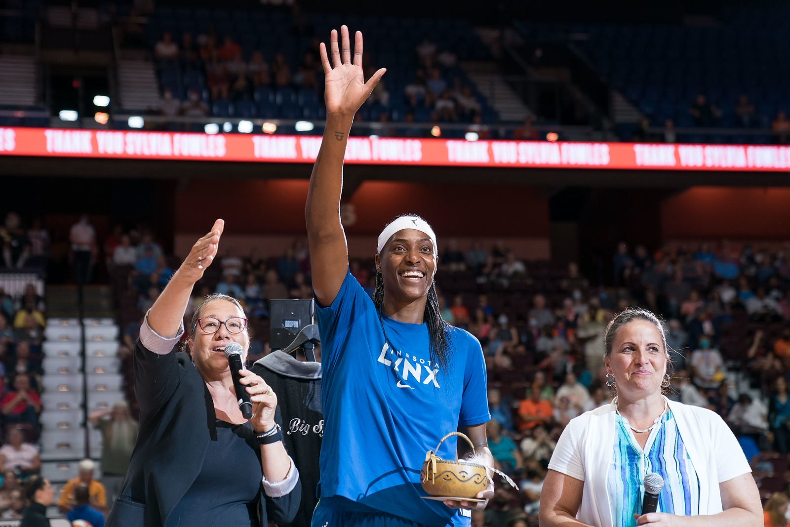 Minnesota Lynx center Sylvia Fowles stands in warmup gear holding her hand up to wave to the crowd while holding a card and small basket in her other hand, as Connecticut Sun President Jennifer Rizzotti stands to her right holding a microphone at chest level, and Mohegan Sun Elder Beth Regan stands to her right, speaking into a handheld microphone and gesturing towards the crowd.