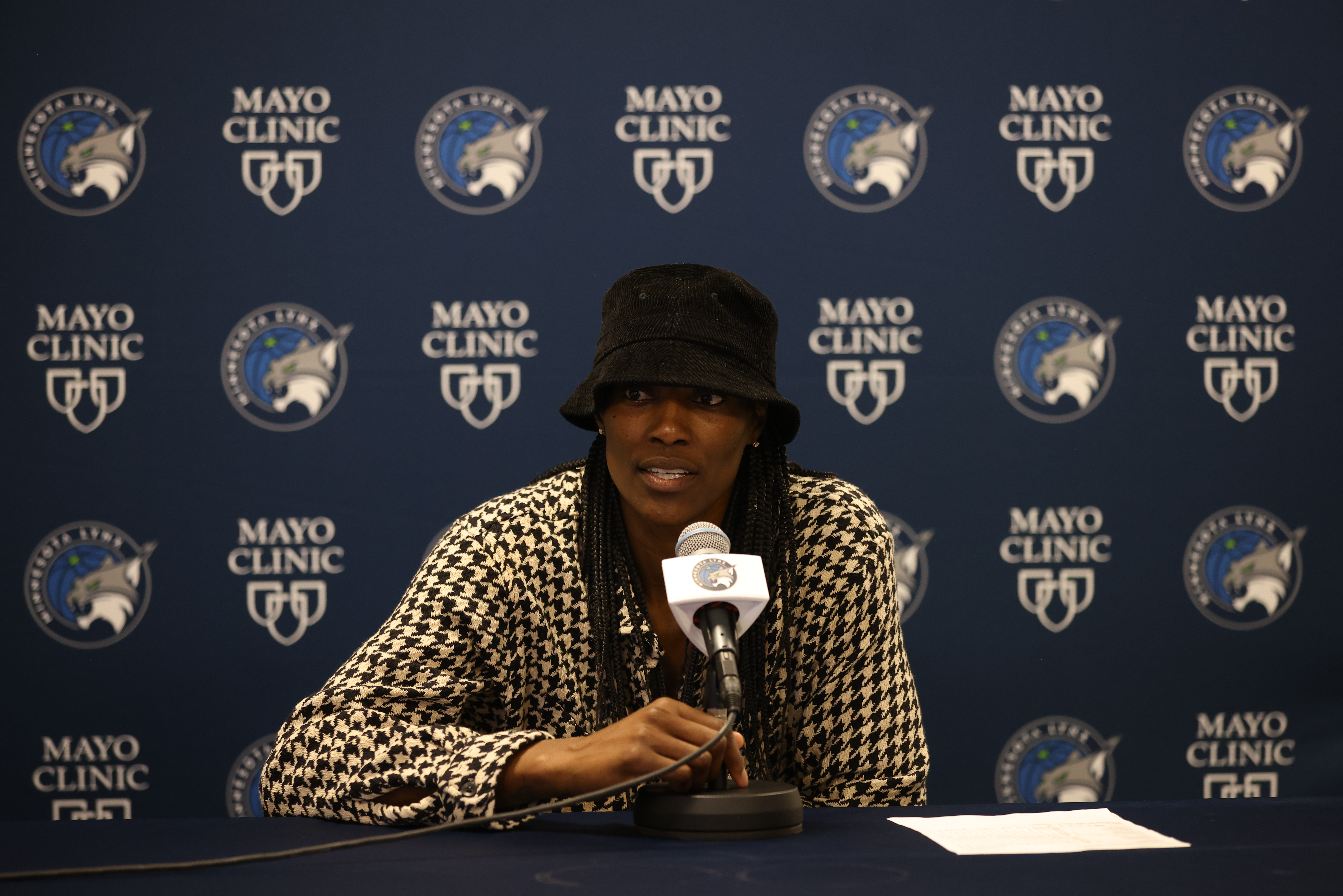 Minnesota center Sylvia Fowles sits behind a table that has a microphone sitting on it directed at her face, wearing a black-and-white-patterned blouse and a black bucket hat during the Lynx's exit interviews.