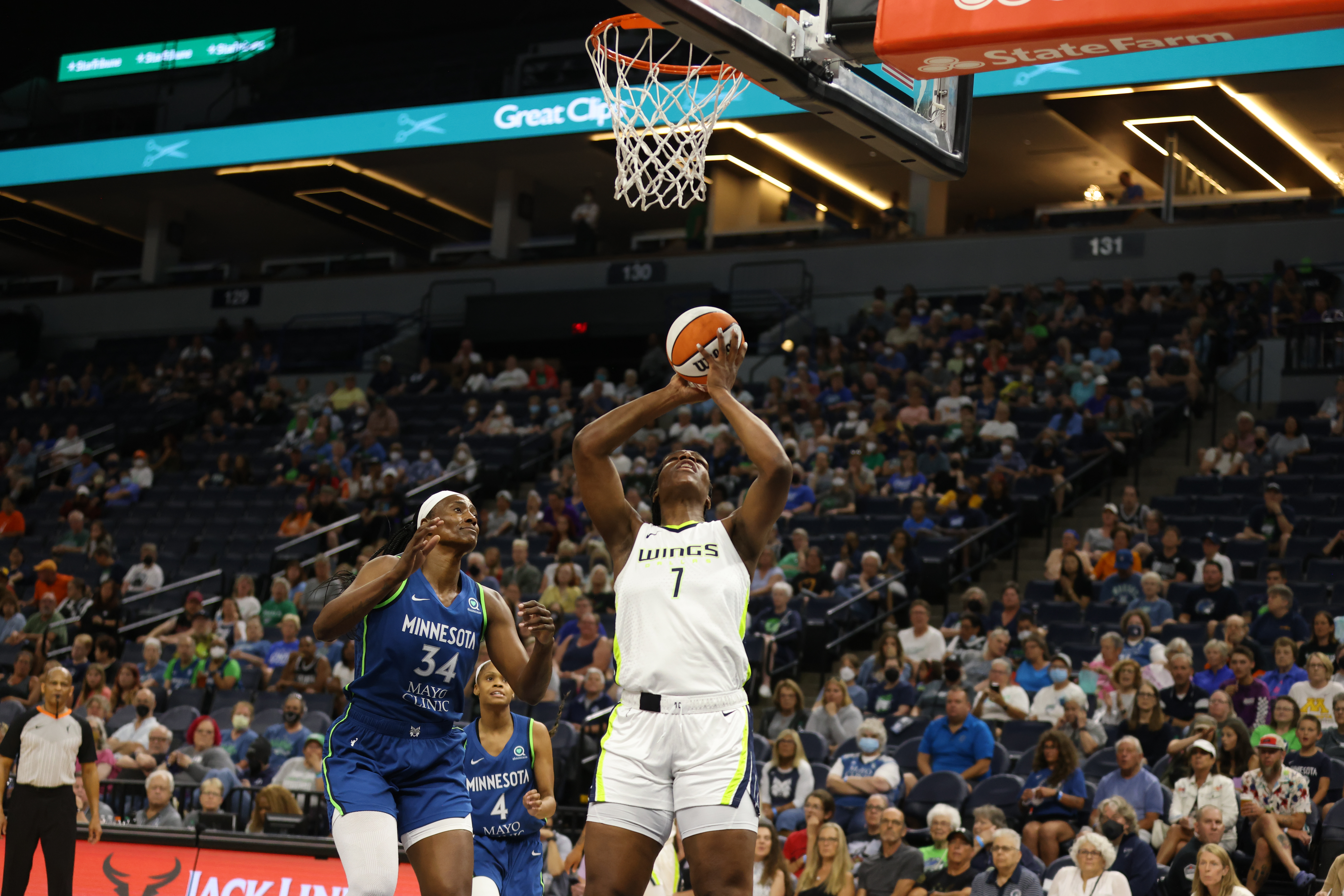 Dallas Wings center Teaira McCowan holds the ball above her head as she stands under the rim looking straight up at the basket, while Minnesota Lynx center Sylvia Fowles stands a foot away, readying to jump towards McCowan and try to block a layup attempt.