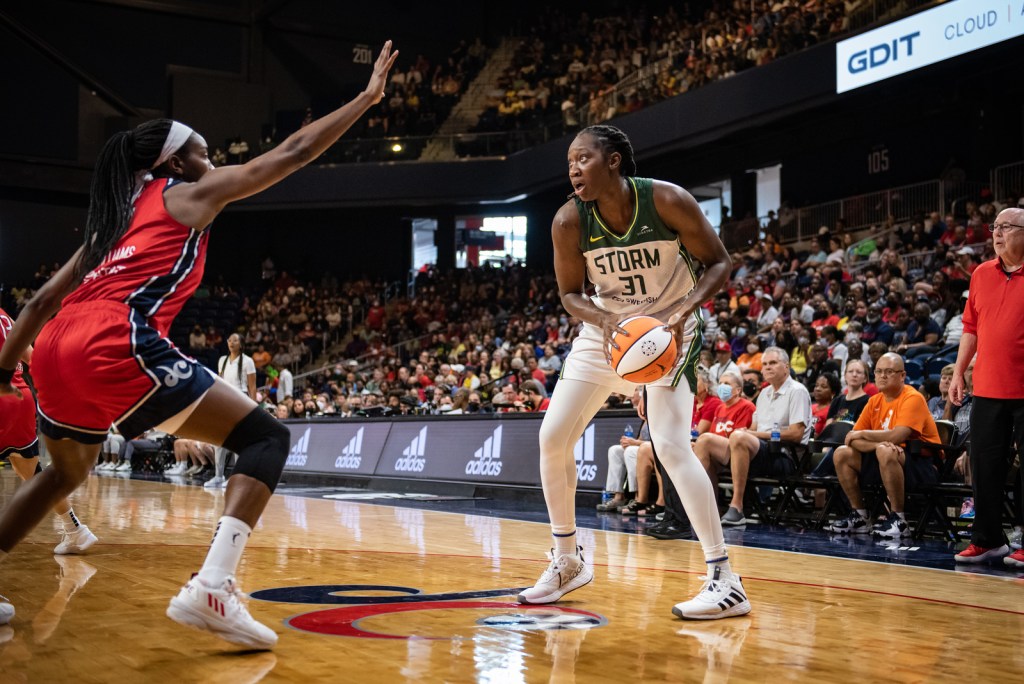 Seattle Storm center Tina Charles holds the ball in a triple-threat stance just inside the left corner as she stares at the hoop, while Washington Mystics center Elizabeth Williams holds a defensive stance from a couple feet in front of her with an arm outstretched towards Charles to prevent a pull-up shot.